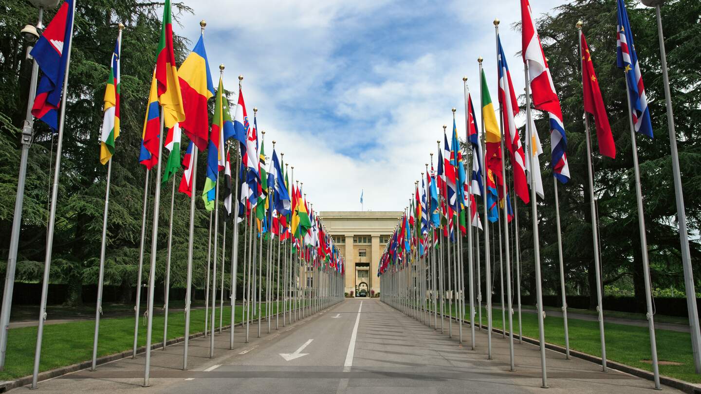 Eingang und Gebäude der Vereinten Nationen in Genf mit Flaggen entlang des Weges | © Gettyimages.com/sumnersgraphicsinc