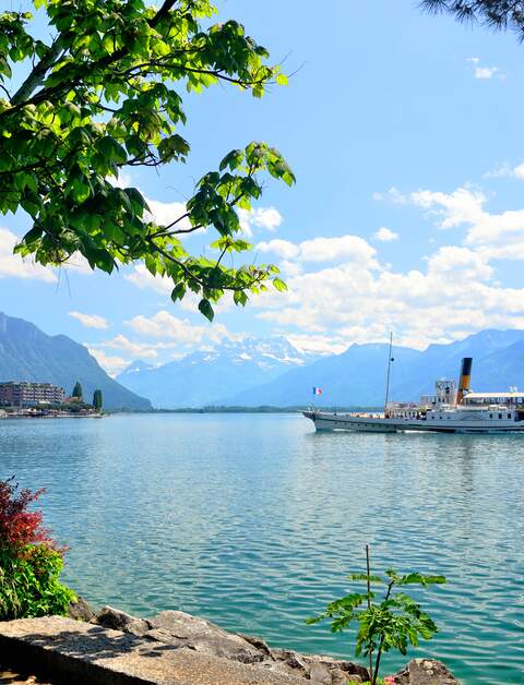 Blick von Montreux auf den Genfersee mit vorbeifahrendem Ausflugsschiff bei Sonnenschein und leichten Wolken | © Gettyimages.com/alxpin