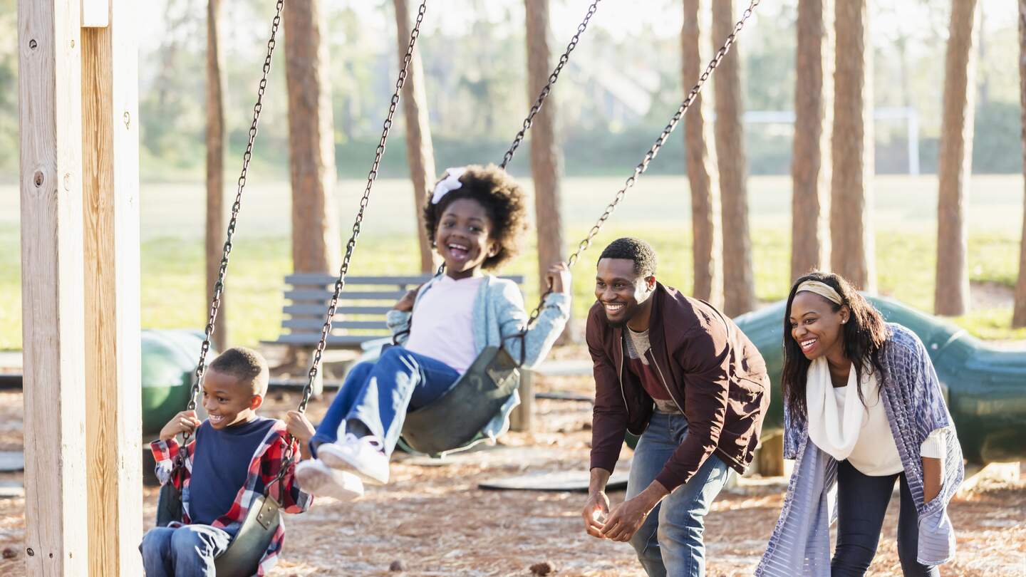 Eine Familie, die zusammen in einem Park spielt und Spaß hat. Die Kinder, ein 7-jähriger Junge und ein 5-jähriges Mädchen, sind auf einer Schaukel und ihre Eltern schubsen ihre Schaukeln an. | © Gettyimages.com/Kali Nine LLC