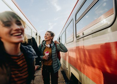 Zwei gut gelaunte, junge Frauen auf dem Bahnsteig zwischen zwei Zügen | © Gettyimages.com/AleksandarNakic