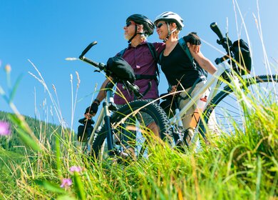 Ein Seniorenpaar steht mit Fahrrädern auf einer grünen Wiese, schaut in die Ferne und ist aus der Untersicht zu sehen, der Himmel ist strahlend blau | © GettyImages.com/nullplus