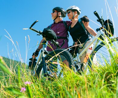 Ein Seniorenpaar steht mit Fahrrädern auf einer grünen Wiese, schaut in die Ferne und ist aus der Untersicht zu sehen, der Himmel ist strahlend blau | © GettyImages.com/nullplus