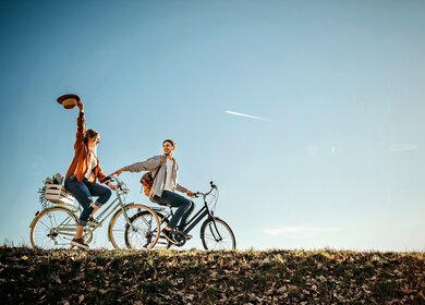 Paar fährt Rad auf dem Deich, die Frau streckt ihre Hand mit Hut in die Höhe und der Mann ist zu ihr gewandt | © Gettyimage/StefaNikolic