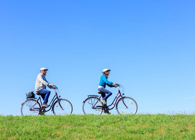 Paar fährt mit dem fahrrad bei blauem Himmel auf strahlend grünem Deich  | © Gettyimage/FredFroese