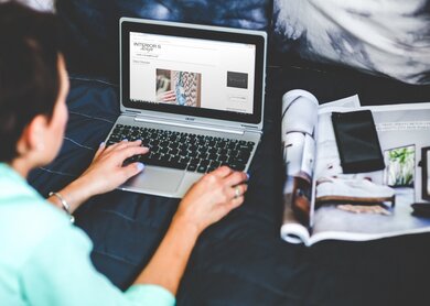Eine Frau, von hinten sichtbar, arbeitet mit ihrem Laptop auf einem Bett und hat einen aufgeschlagenen Katalog und ihr Handy neben sich liegen | © Pixabay/kaboompics