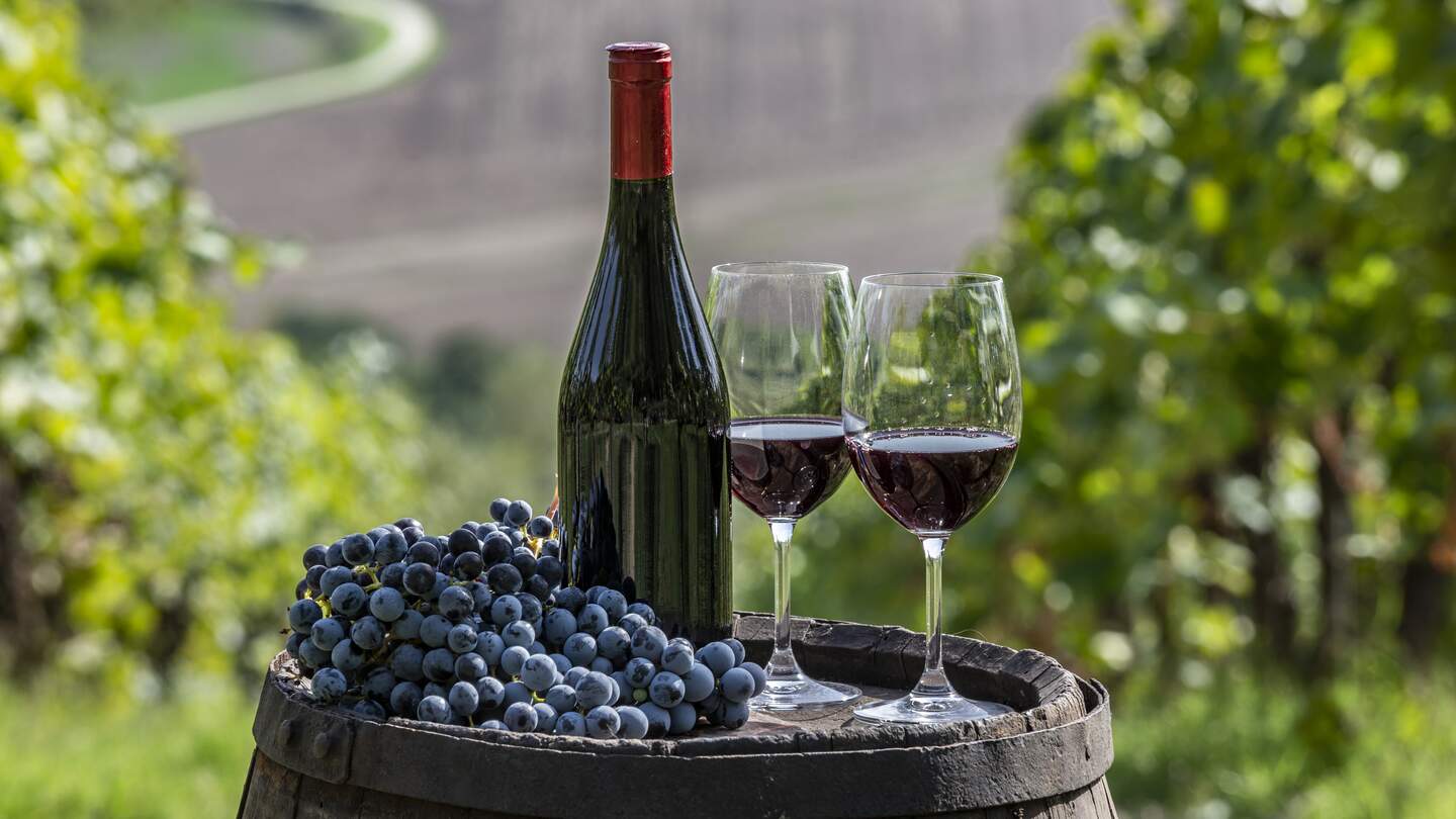 Fass auf dem eine Flasche Rotwein und zwei gefüllte Rotwein Gläser stehen, daneben liegen Trauben und im Hintergrund sind Weinberge zu sehen | © Gettyimages.com/Alberto Gagliardi